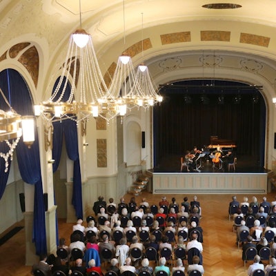 Menschen sitzen auf Stühlen in einem festlichen Saal mit großen Kronleuchtern; auf der Bühne spielt ein Orchester.