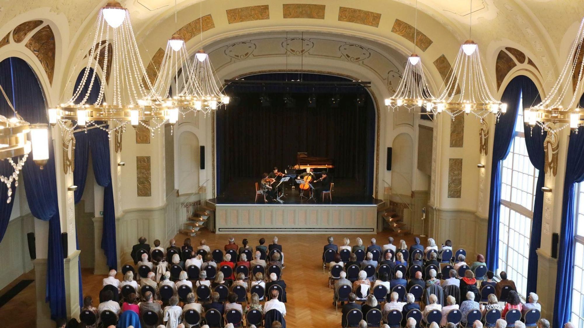 Menschen sitzen auf Stühlen in einem festlichen Saal mit großen Kronleuchtern; auf der Bühne spielt ein Orchester.