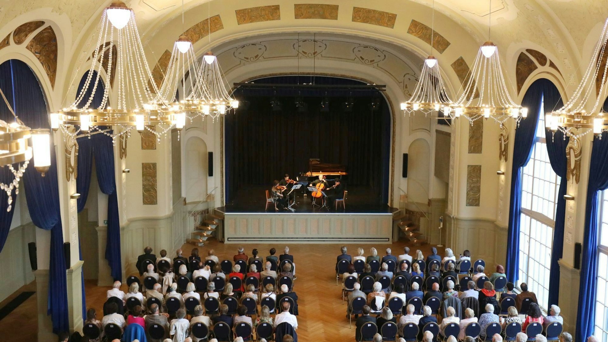 Menschen sitzen auf Stühlen in einem festlichen Saal mit großen Kronleuchtern; auf der Bühne spielt ein Orchester.