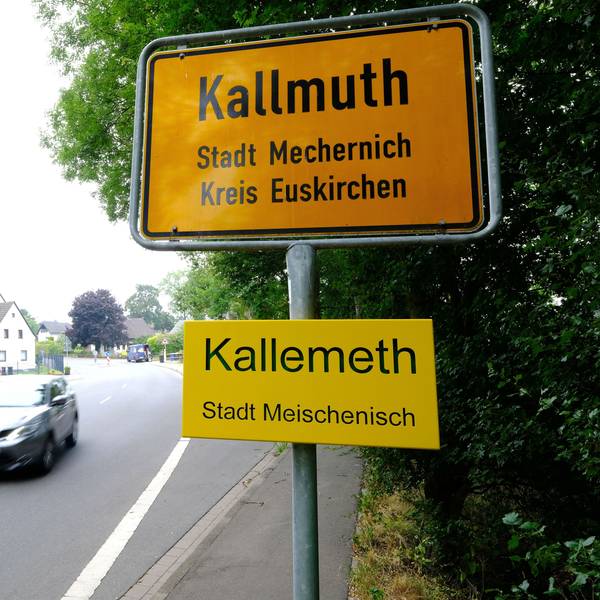 Ortstafel des Mechernicher Stadtteils Kallmuth mit Zusatzschild des Ortsnamens in Eifeler Mundart („Kallemeth, Stadt Meischenisch“). Auf der Dorfstraße fährt ein Auto vorbei.