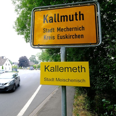 Ortstafel des Mechernicher Stadtteils Kallmuth mit Zusatzschild des Ortsnamens in Eifeler Mundart („Kallemeth, Stadt Meischenisch“). Auf der Dorfstraße fährt ein Auto vorbei.