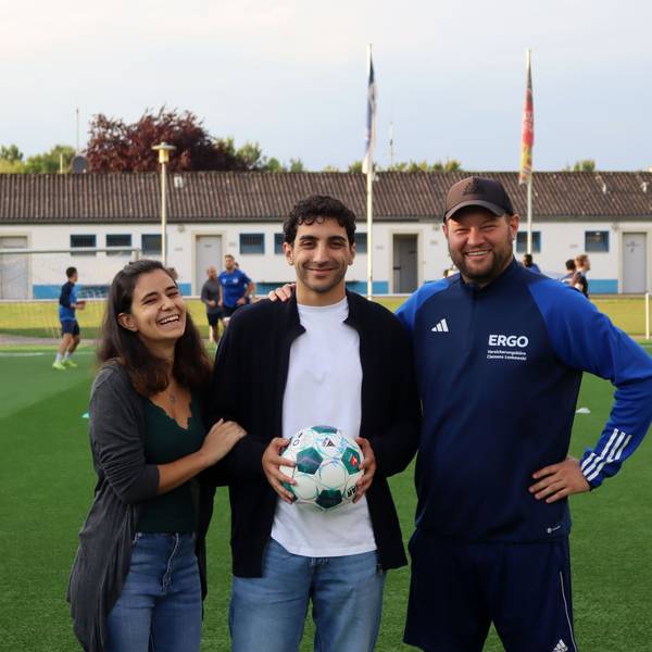 Patrick Hinsch hält einen Ball in den Händen. Seine Freundin Daria Scala hat sich eingehakt, sein Trainer Frederik Ziburske legt ihm die Hand auf die Schulter. Im Hintergrund trainert der SSV Weilerswist.