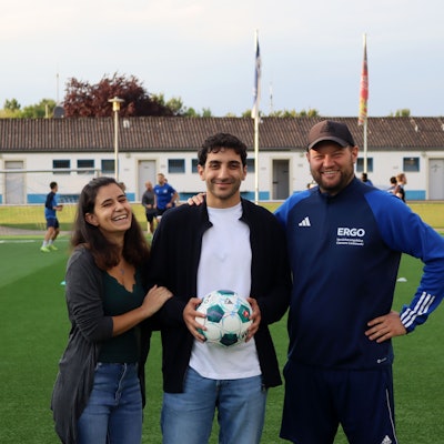 Patrick Hinsch hält einen Ball in den Händen. Seine Freundin Daria Scala hat sich eingehakt, sein Trainer Frederik Ziburske legt ihm die Hand auf die Schulter. Im Hintergrund trainert der SSV Weilerswist.