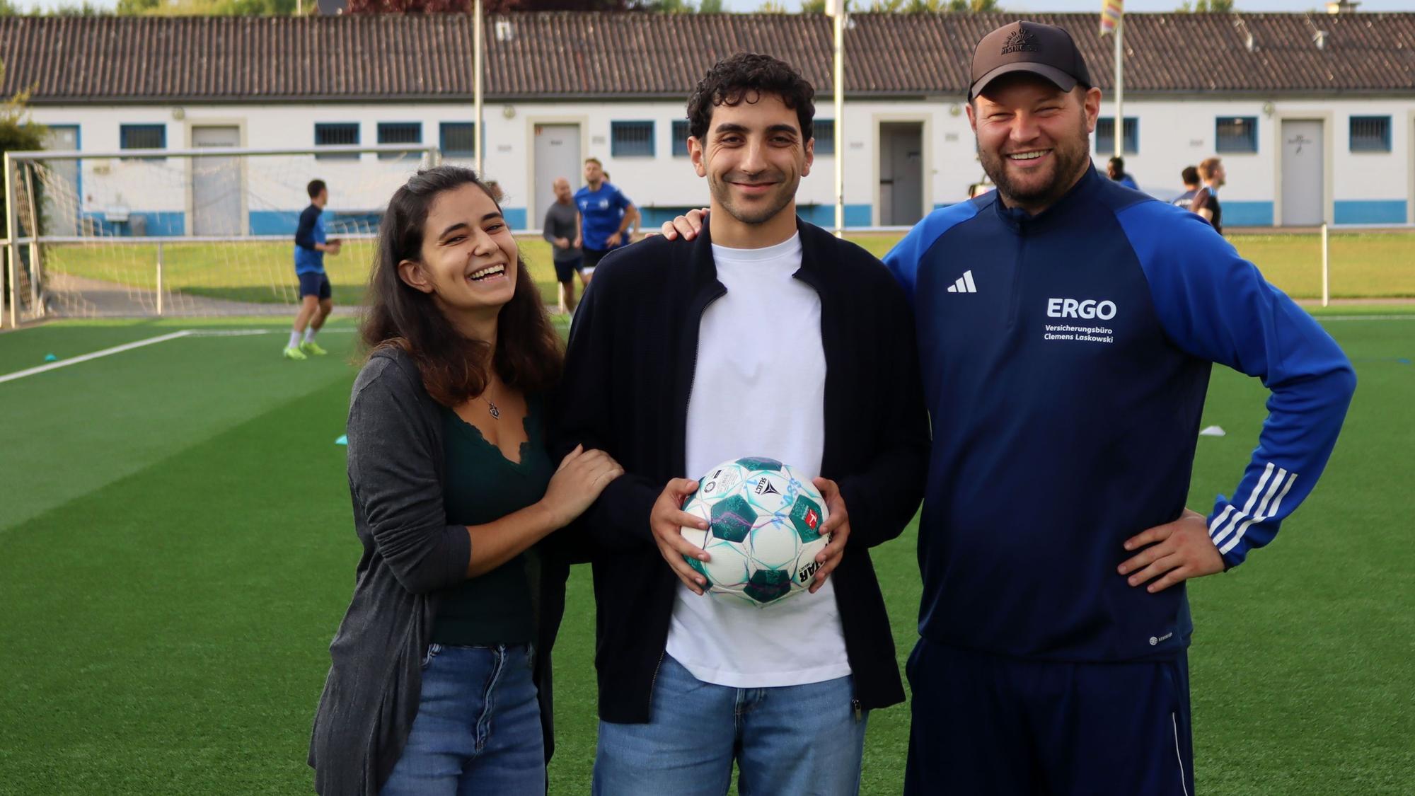 Patrick Hinsch hält einen Ball in den Händen. Seine Freundin Daria Scala hat sich eingehakt, sein Trainer Frederik Ziburske legt ihm die Hand auf die Schulter. Im Hintergrund trainert der SSV Weilerswist.