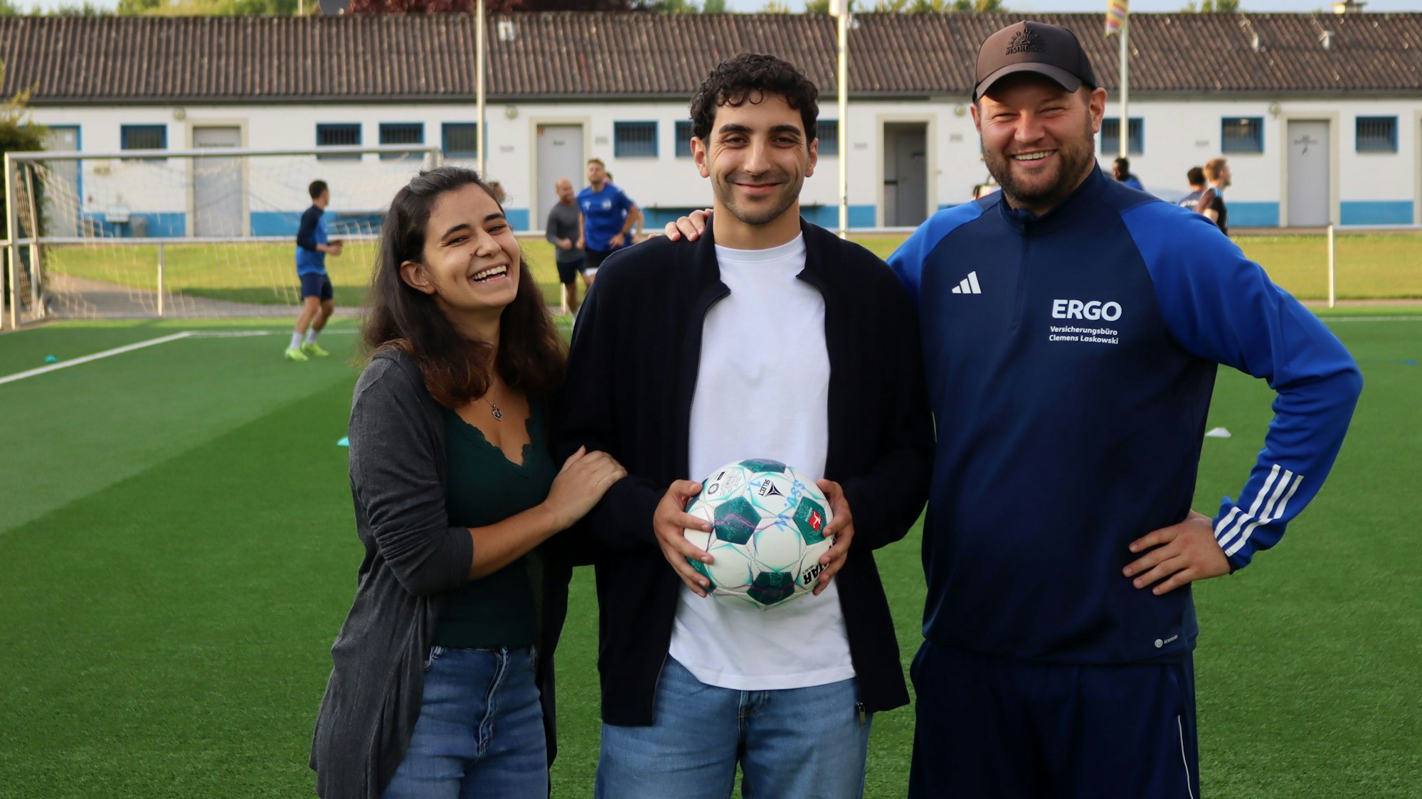 Patrick Hinsch hält einen Ball in den Händen. Seine Freundin Daria Scala hat sich eingehakt, sein Trainer Frederik Ziburske legt ihm die Hand auf die Schulter. Im Hintergrund trainert der SSV Weilerswist.