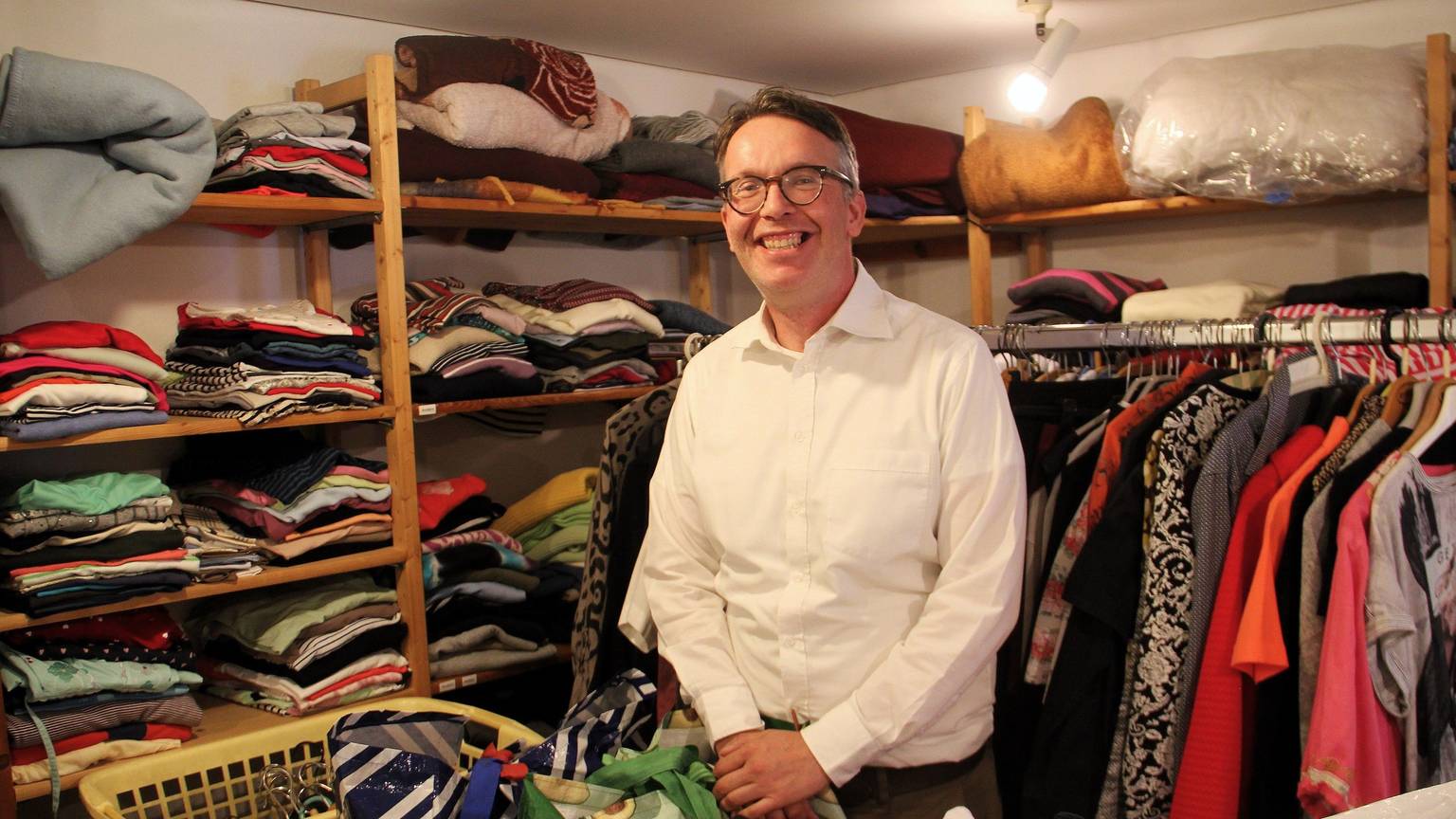 Ein Mann mit grauem Haar und Brille steht inmitten von Regalen und Kleiderstange mit Kleidung.