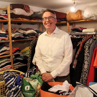 Ein Mann mit grauem Haar und Brille steht inmitten von Regalen und Kleiderstange mit Kleidung.
