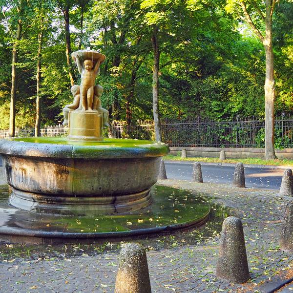 Ein runder Brunnen mit Kinderfiguren in der Mitte ist auf einem kleinen Platz umgeben von Pollern.