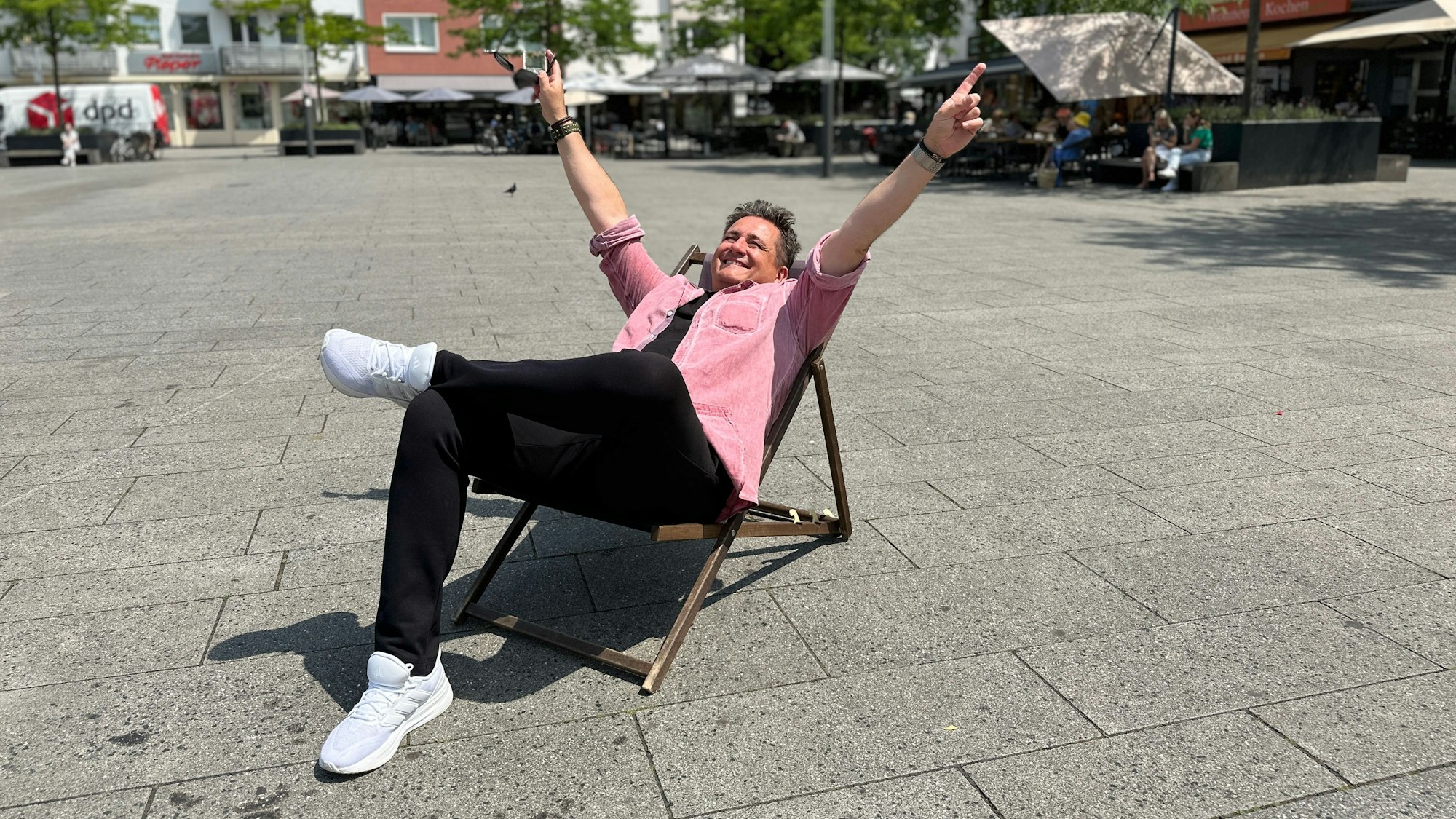 Ein Mann im rosa Hemd und schwarzer Hose sitzt in einem Liegestuhl auf einem großen Platz.
