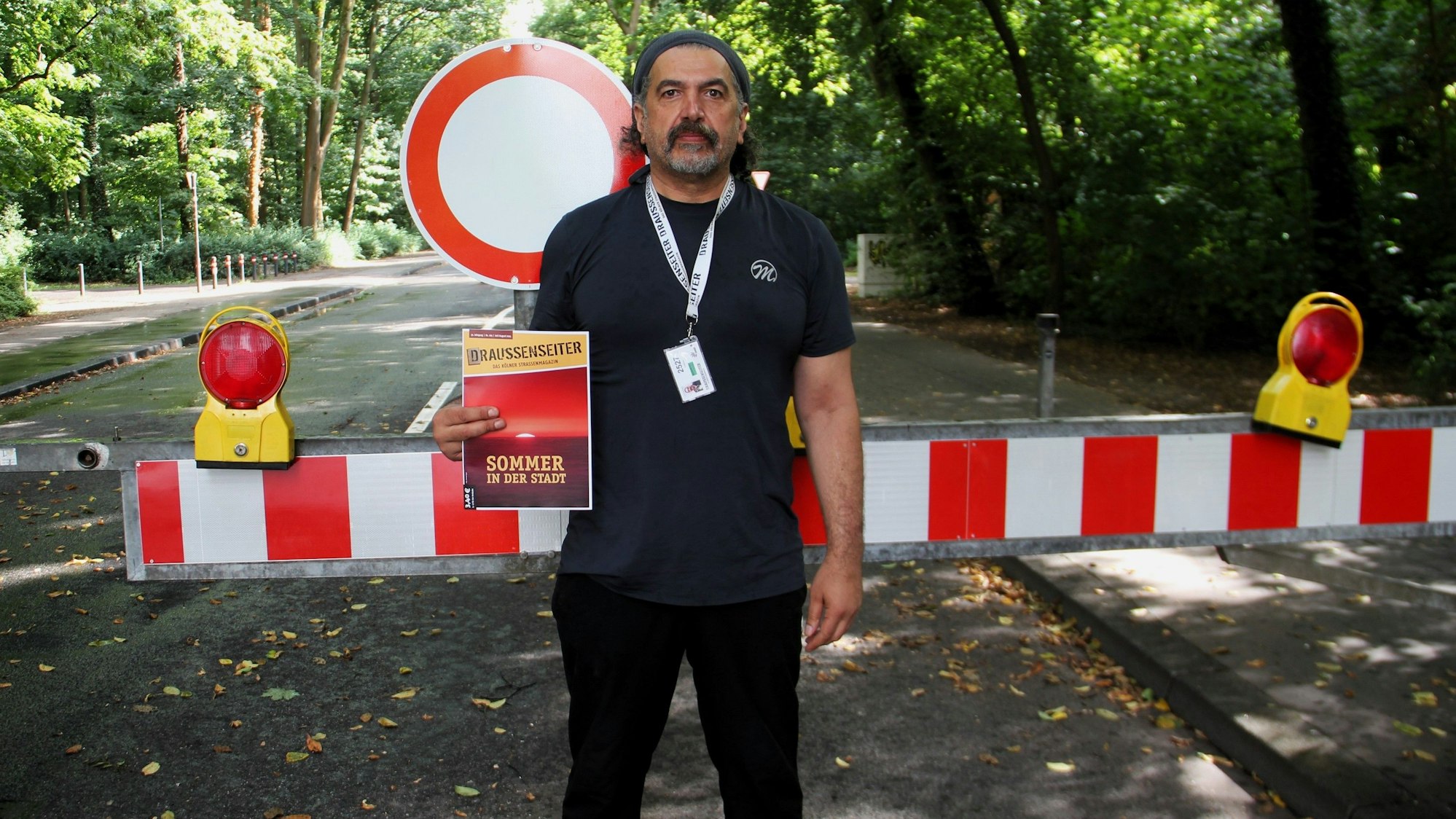 Ein Mann steht vor einer rot-weißen Schranke, die die Zufahrt zu einer Straße verbietet, die in einem Wald liegt.