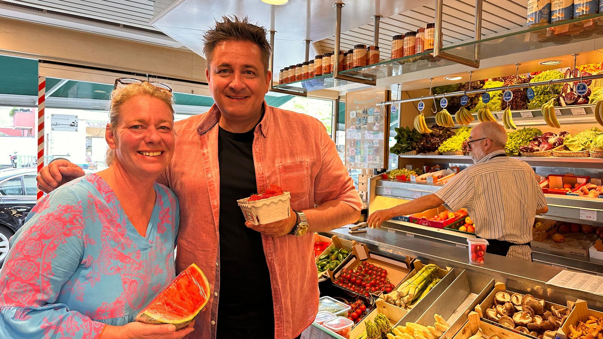 Ein Mann und eine Frau stehen nebeneinander in einem Obst- und Gemüse-Geschäft. Sie hält eine Viertel Wassermelone, er eine Schale Erdbeeren.