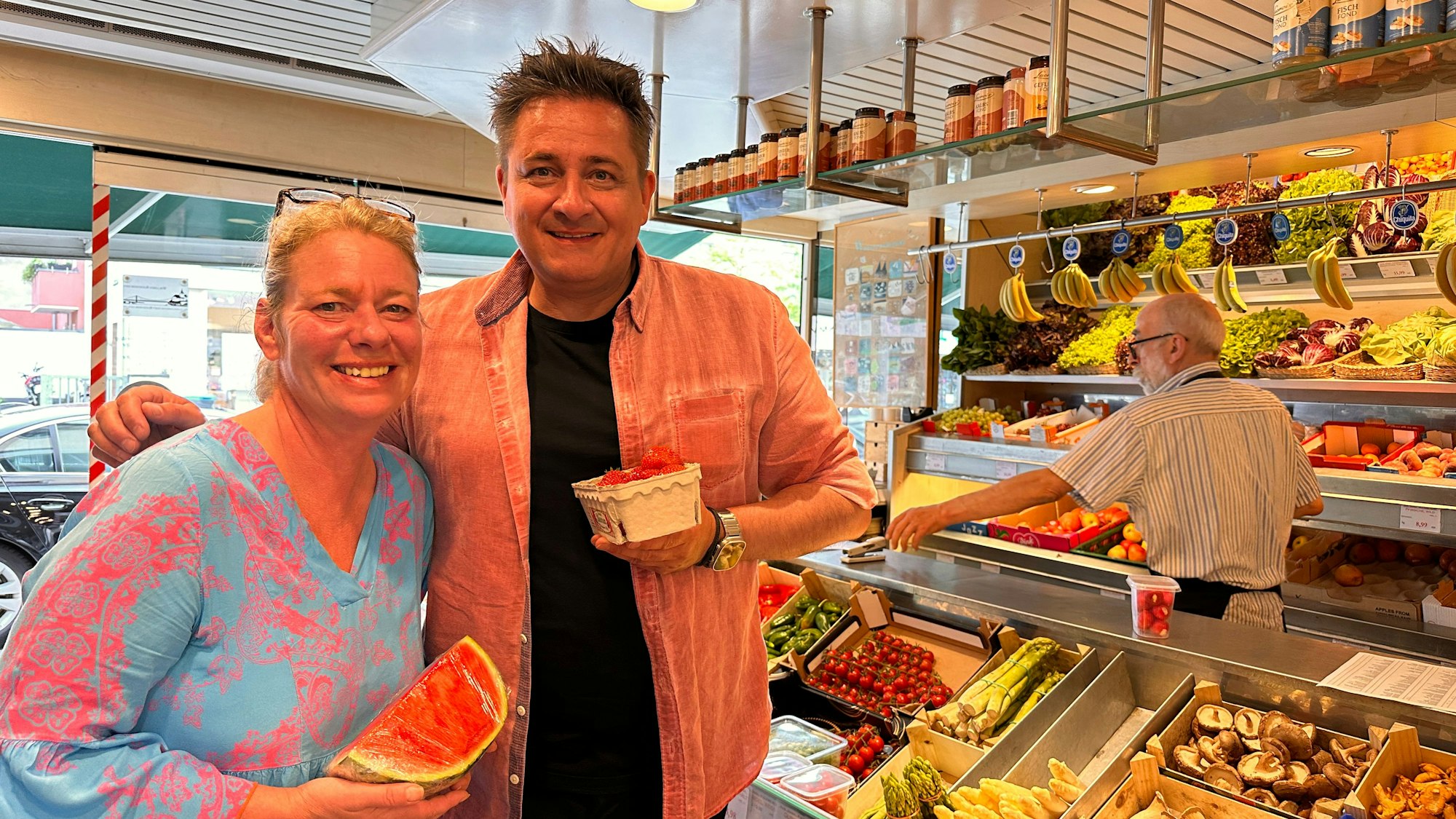 Ein Mann und eine Frau stehen nebeneinander in einem Obst- und Gemüse-Geschäft. Sie hält eine Viertel Wassermelone, er eine Schale Erdbeeren.