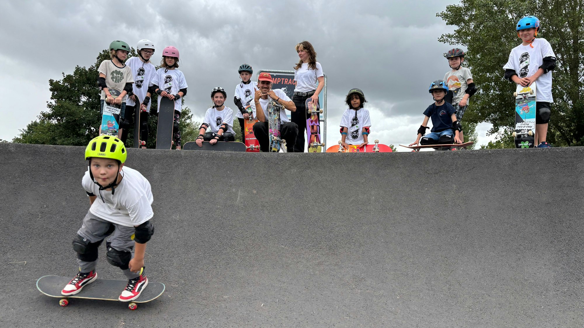 Ein Junge mit Helm und Schützern fährt auf einem Skateboard in eine Kurve des Weilerswister Pumptracks. Im Hintergrund sind andere Kinder, ein Betreuer und eine Betreuerin.