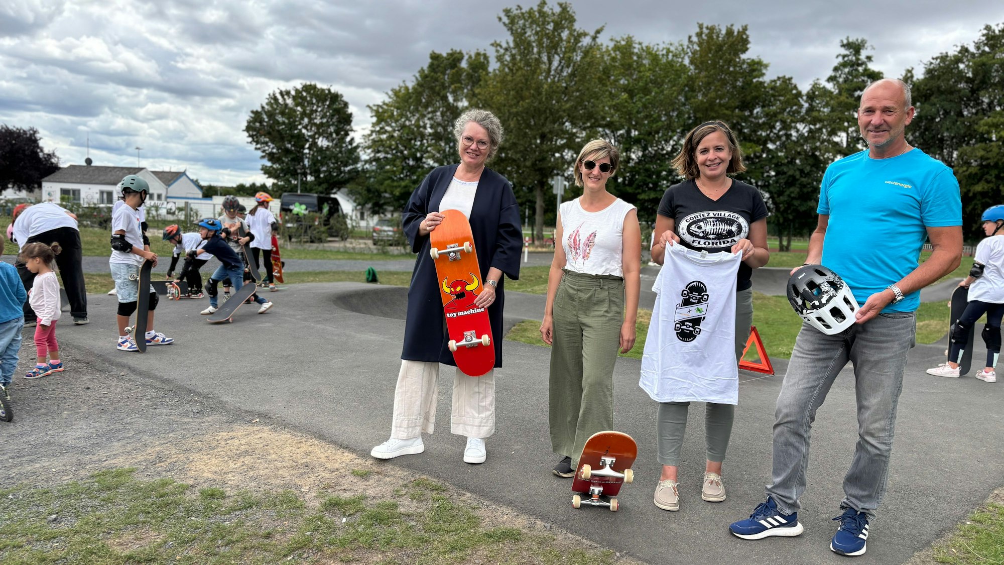 Anne Horst, Kerri Wetter, Stefanie Klein und Achim Diewald stehen auf dem Weilerswister Pumptrack. Die Bürgermeisterin hält ein Skateboard. Im Hintergrund stehen Kinder und Jugendliche mit Skateboards.