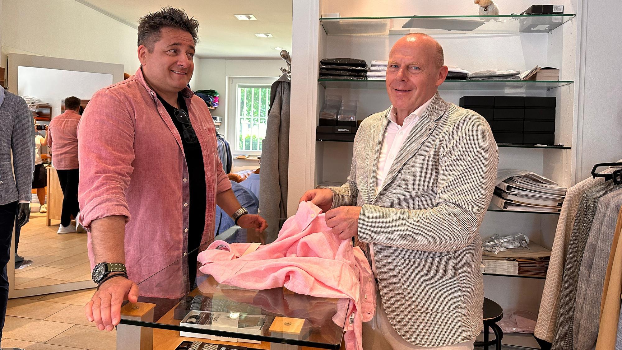 Ein Mann im rosa Hemd steht in einem Herrenmode-Geschäft, der Inhaber des Geschäfts berät ihn.
