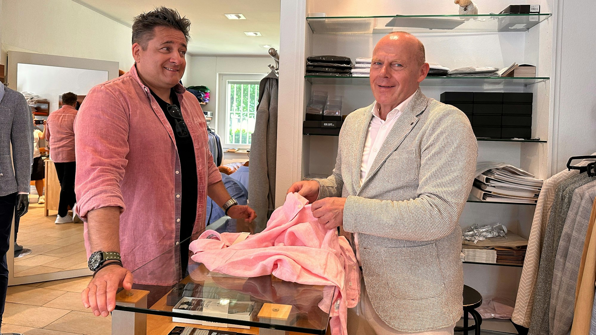 Ein Mann im rosa Hemd steht in einem Herrenmode-Geschäft, der Inhaber des Geschäfts berät ihn.