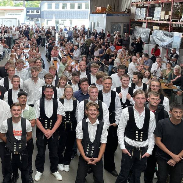 Alle 33 neuen Experten im Dachdecker- und Baugewerkshandwerk stehen für das Gruppenfoto parat. Dahinter haben sich Freunde und Familie auf Bierbänken versammelt.
