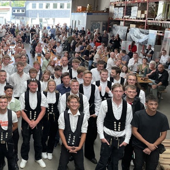 Alle 33 neuen Experten im Dachdecker- und Baugewerkshandwerk stehen für das Gruppenfoto parat. Dahinter haben sich Freunde und Familie auf Bierbänken versammelt.