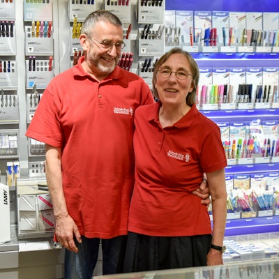 Eine Frau und ein Mann in roten Polohemden stehen vor einer Wand mit Stiften.