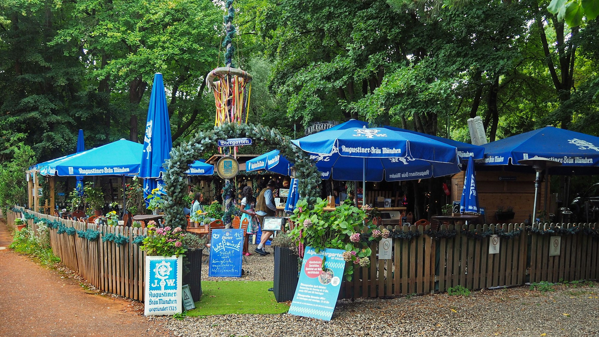 Ein mit einem hüfthohen Holzzaun umzäunter Biergarten ist zu sehen, der viel blau-weiße Deko hat und so bayrisches Ambiente verströmt.