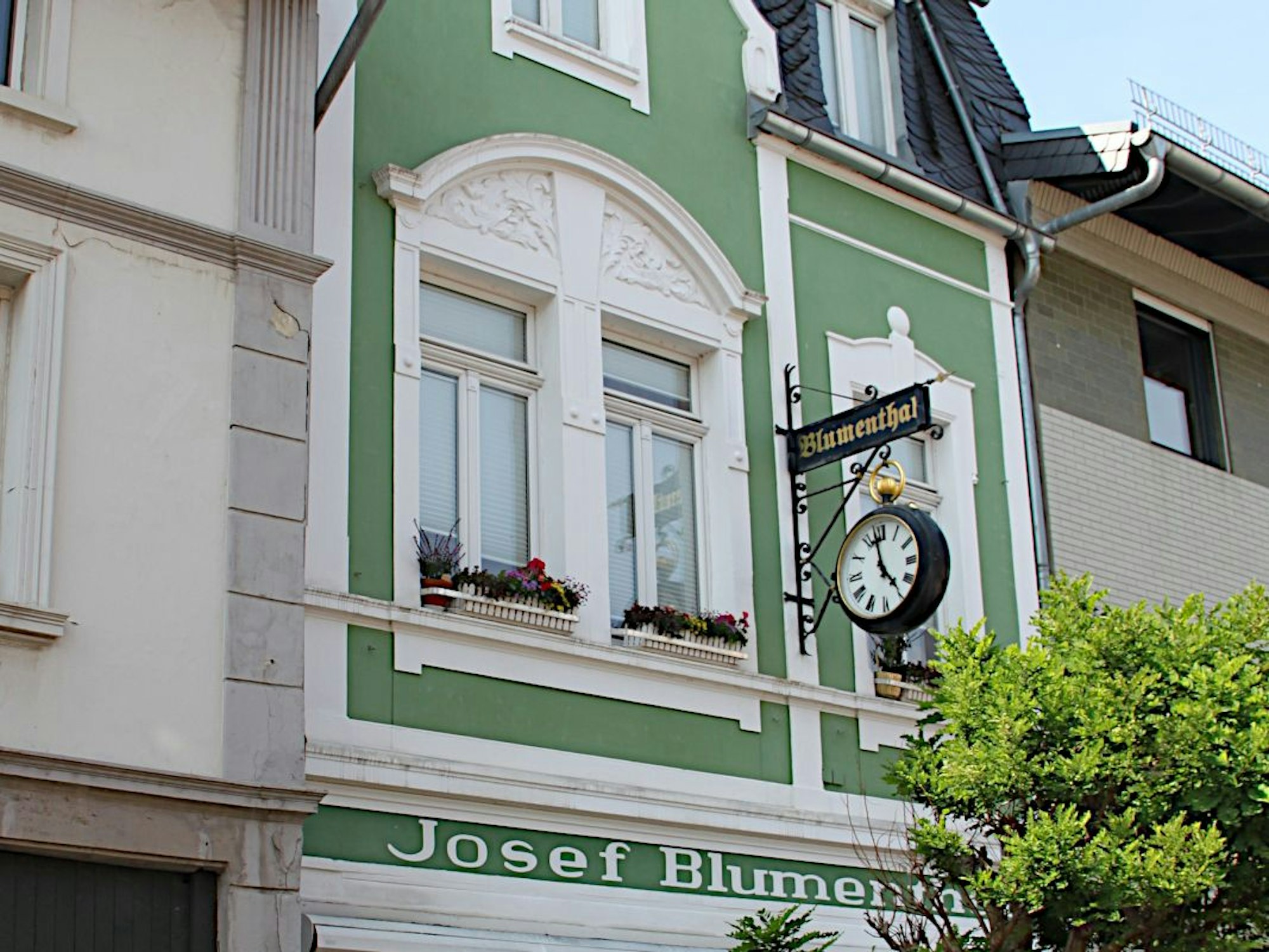 Aufnahme der Fassade des Fachgeschäfts von Josef Blumenthal. Vor der grün-weißen Fassade hängt eine Uhr.