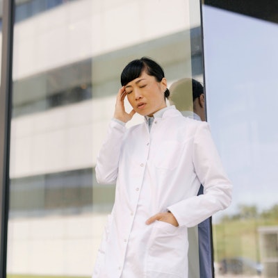 Eine Ärztin steht vor einer Glaswand und hält sich angestrengt den Kopf.