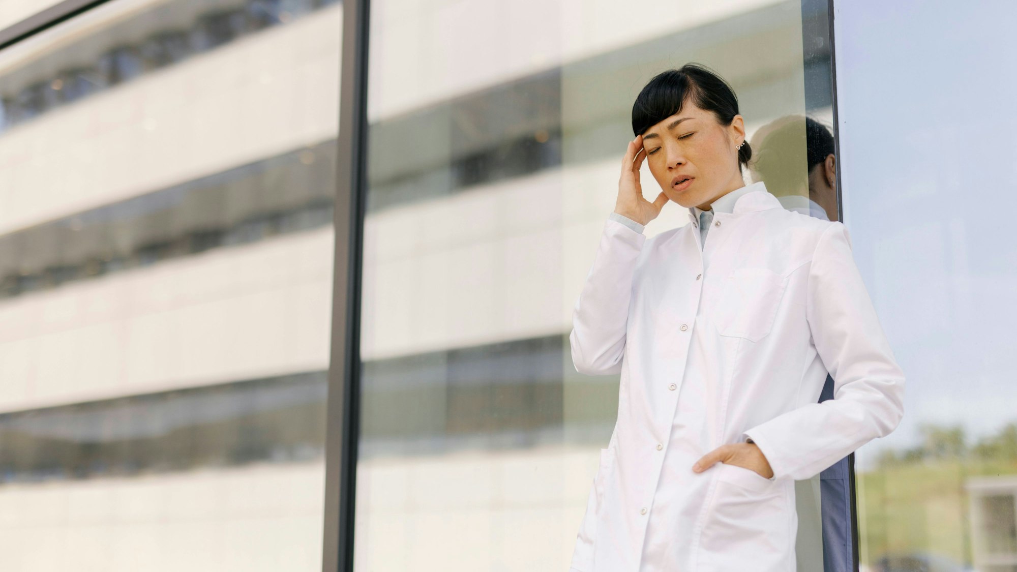 Eine Ärztin steht vor einer Glaswand und hält sich angestrengt den Kopf.