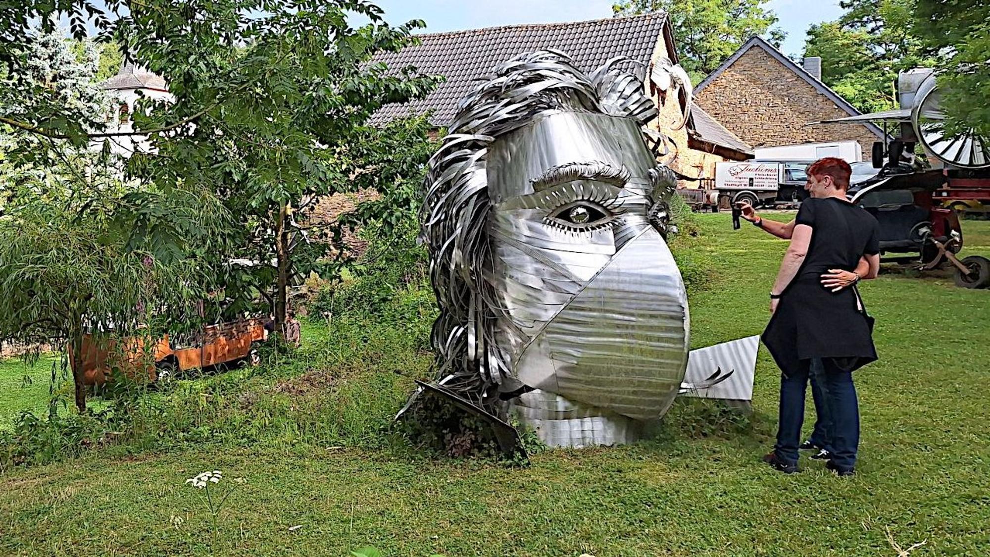 Eine große silberne Metallskulptur, die Beethovens Gesichtszüge erahnen lässt, steht auf der Wiese in einem Garten. Eine Frau steht davor und betrachtet das Werk.