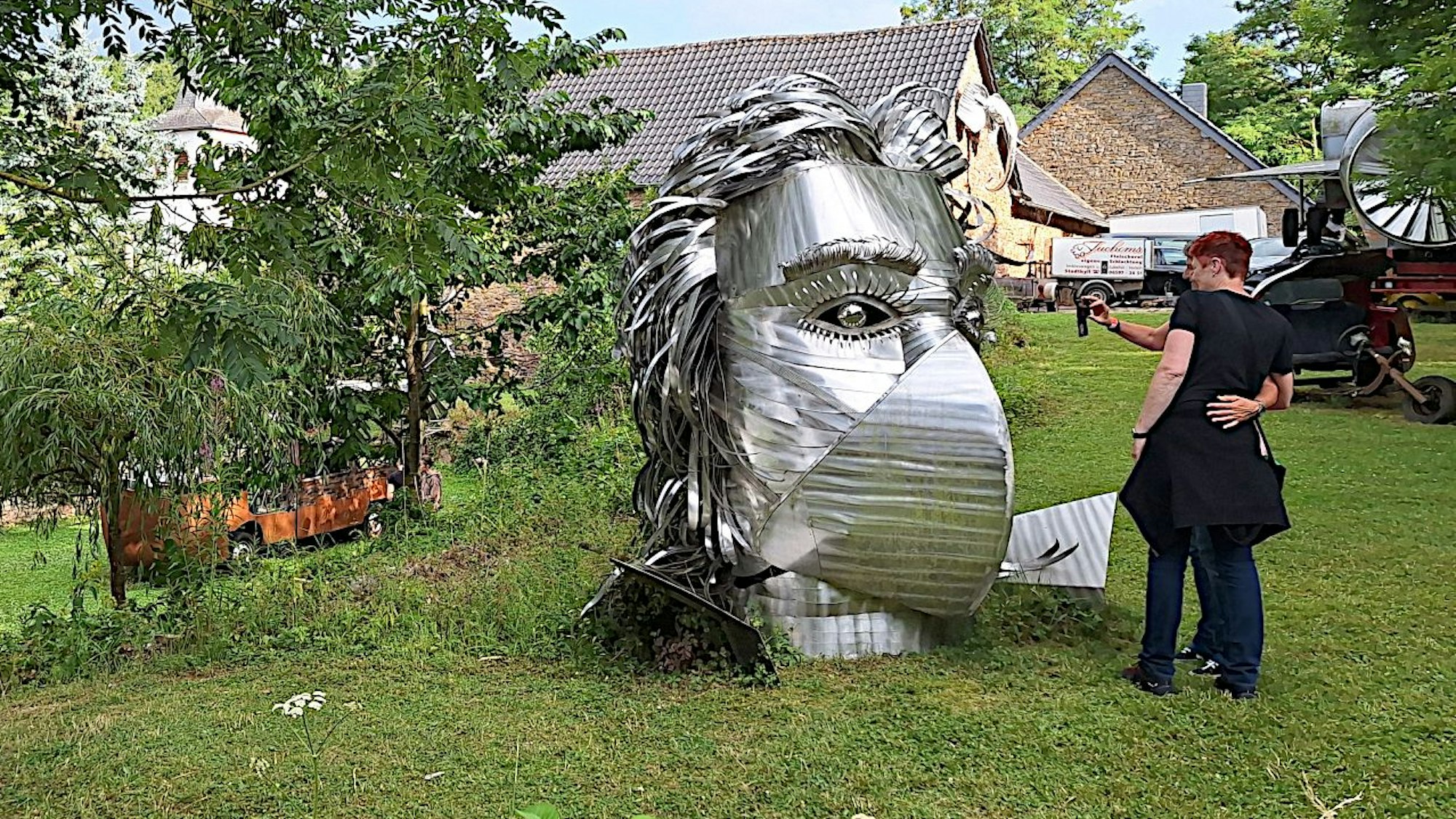 Eine große silberne Metallskulptur, die Beethovens Gesichtszüge erahnen lässt, steht auf der Wiese in einem Garten. Eine Frau steht davor und betrachtet das Werk.