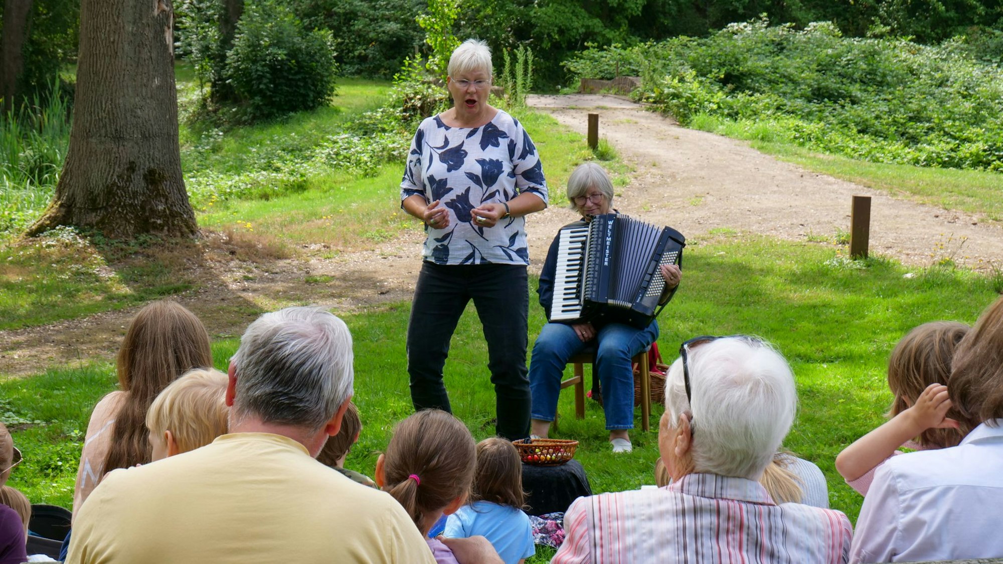 Vor einer Gruppe mit Menschen im grünen Park steht Angersbach und erzählt eine Geschichte. Neben ihr sitzt Mecki Claus auf einem Stuhl und spielt Akkordeon.