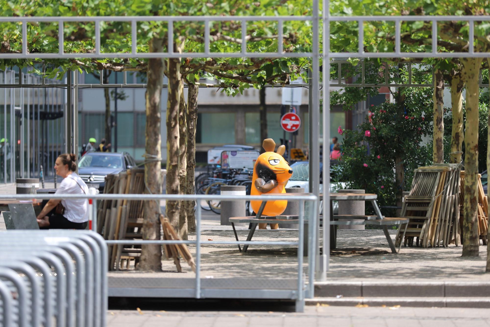 Mit einem großen Brandfleck steht die Maus-Figur hinter einer Sitzfläche vor dem WDR-Gebäude.