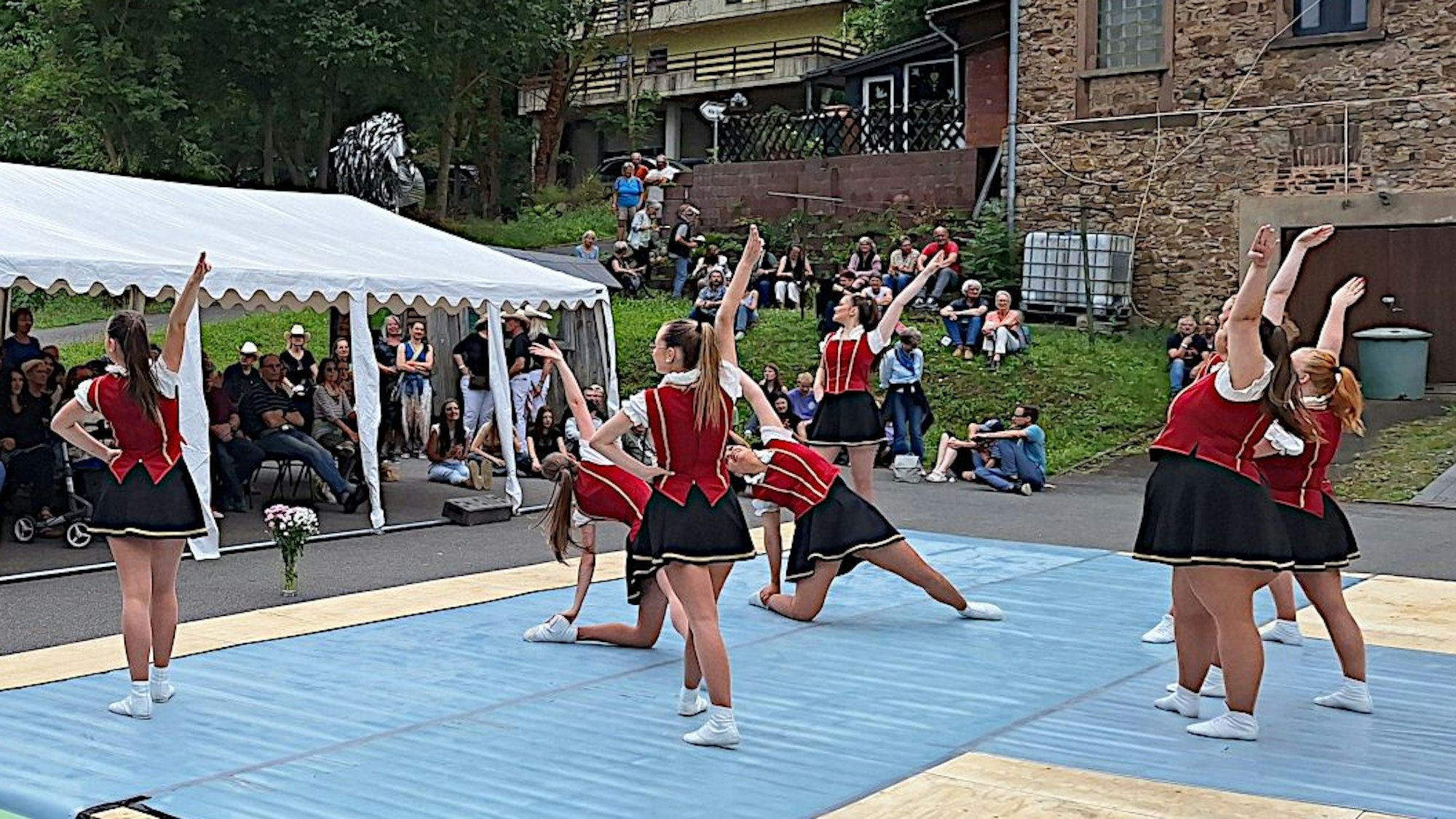 Gardetänzerinnen in rot-schwarzen Kostümen tanzen auf blauen Matten vor Publikum, das auf der Wiese und unter einem Zeltpavillon sitzt.