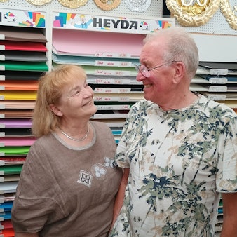 Das Ehepaar schaut sich an und lächelt einander zu. Im Hintergrund liegt reihenweise buntes Papier in Regalen aus.