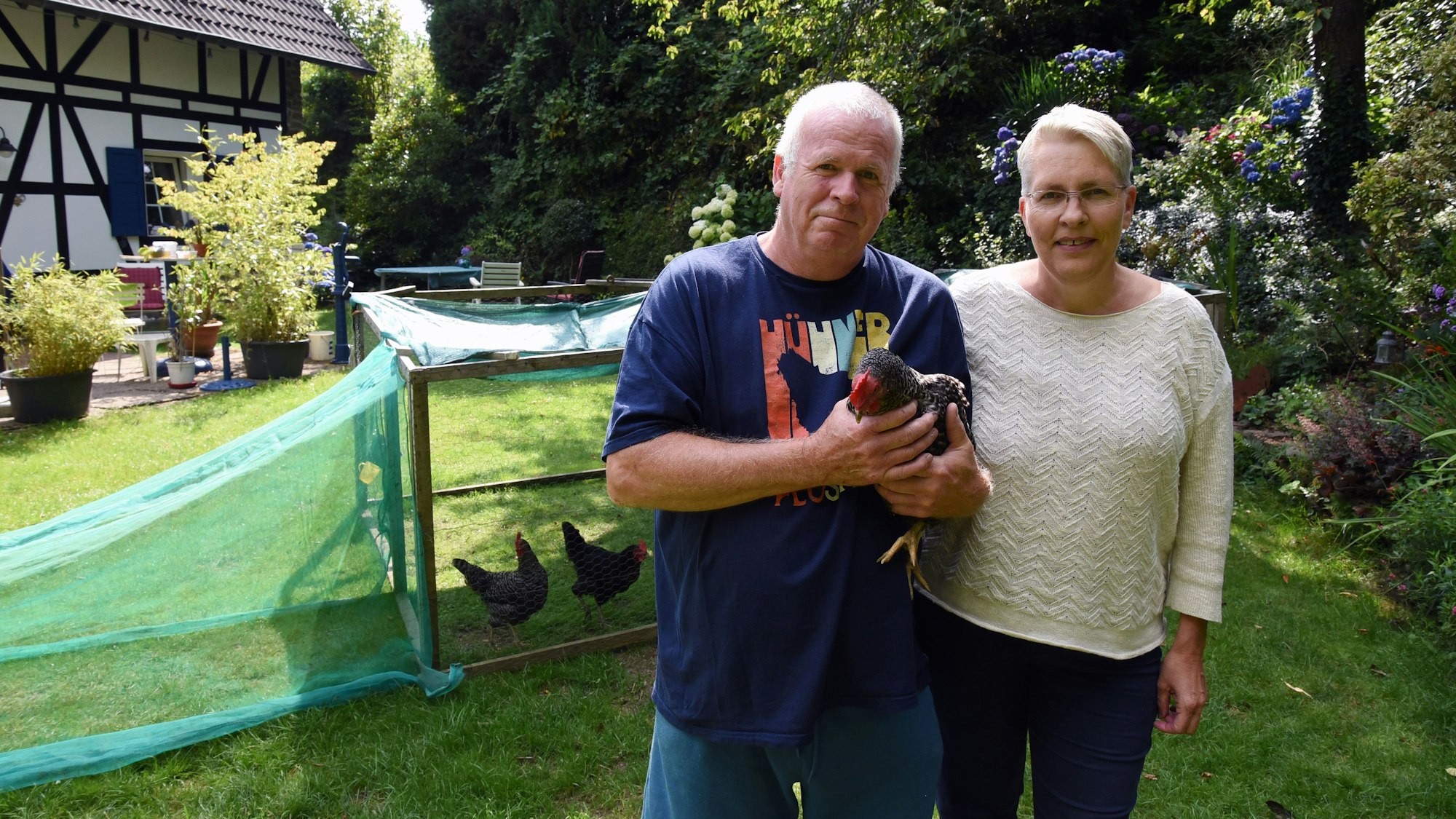 Ein Ehepaar steht in ihrem Garten vor einem Hühnerstall. Er hällt ein Huhn in den Händen.