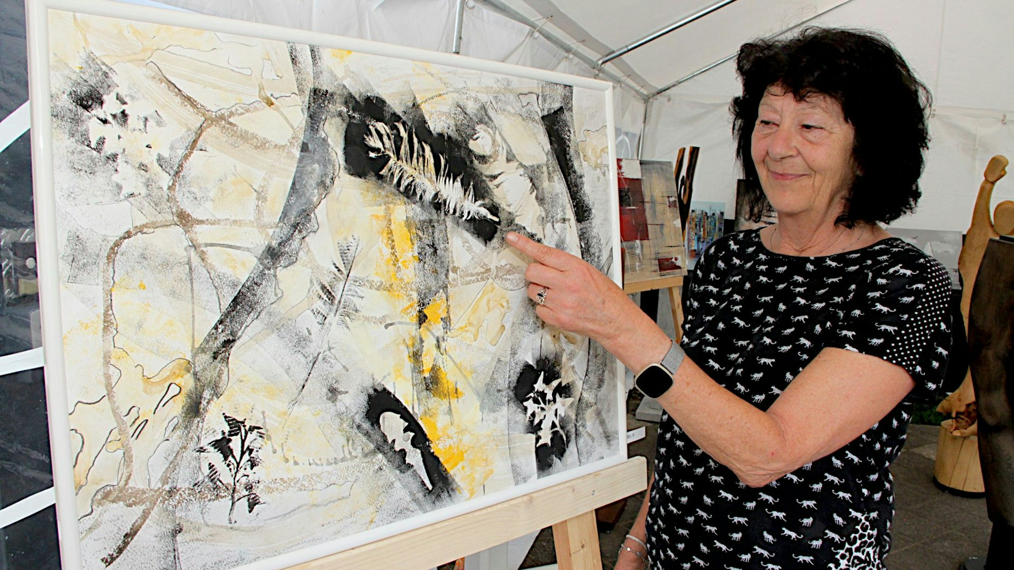 Eine Künstlerin steht neben einem Gemälde, auf das sie blickt und mit dem linken Zeigefinger zeigt.