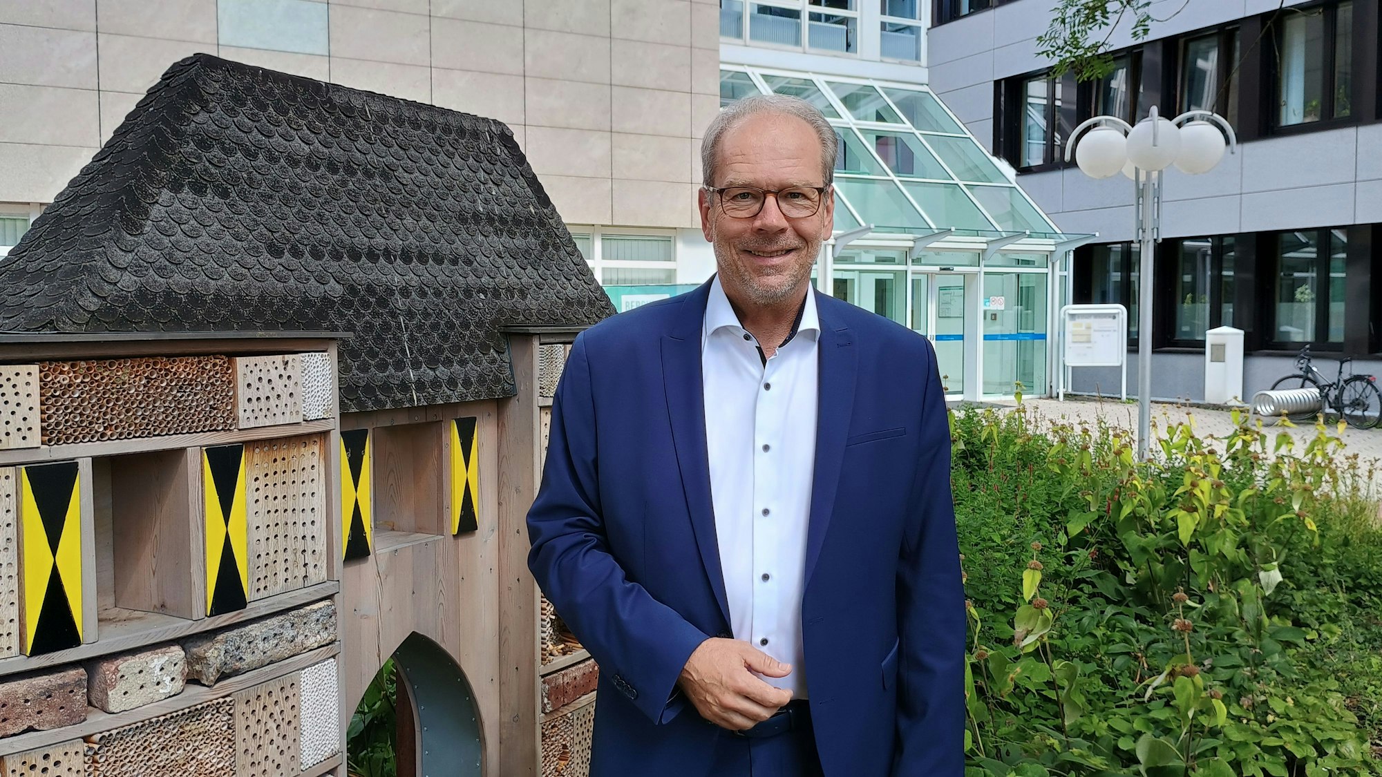 Volker Mießeler trägt ein blaues Sakko und ein helles Hemd. Er steht vor dem Rathaus in Bergheim, rechts neben sich ein Insektenhotel, das wie das Aachener Tor aussieht.
