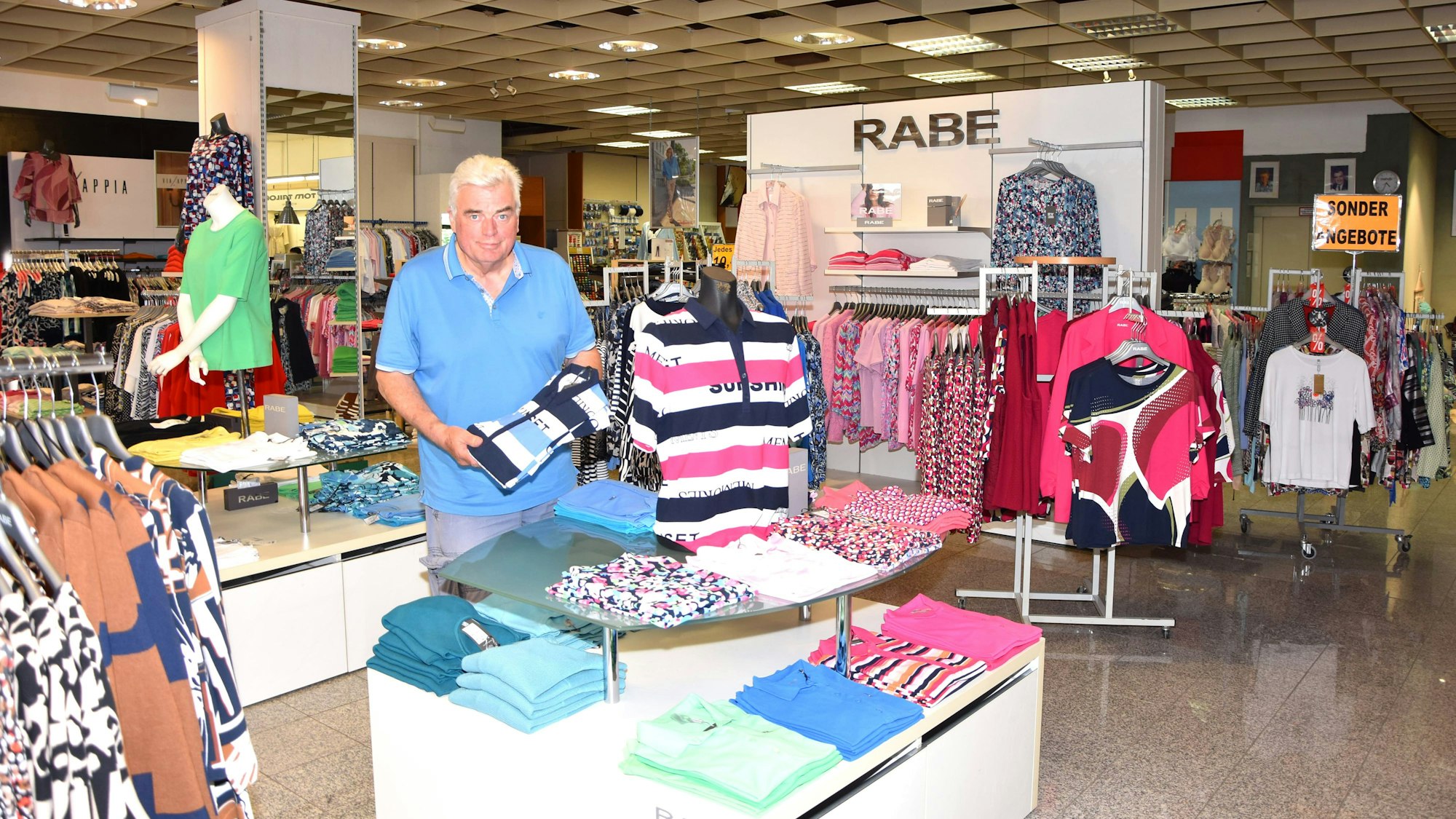 Hermann-Josef Peters hält einen Pullover. Er steht in seinem Laden, es liegen Hemden aus und Blusen hängen an Kleiderständerregalen.