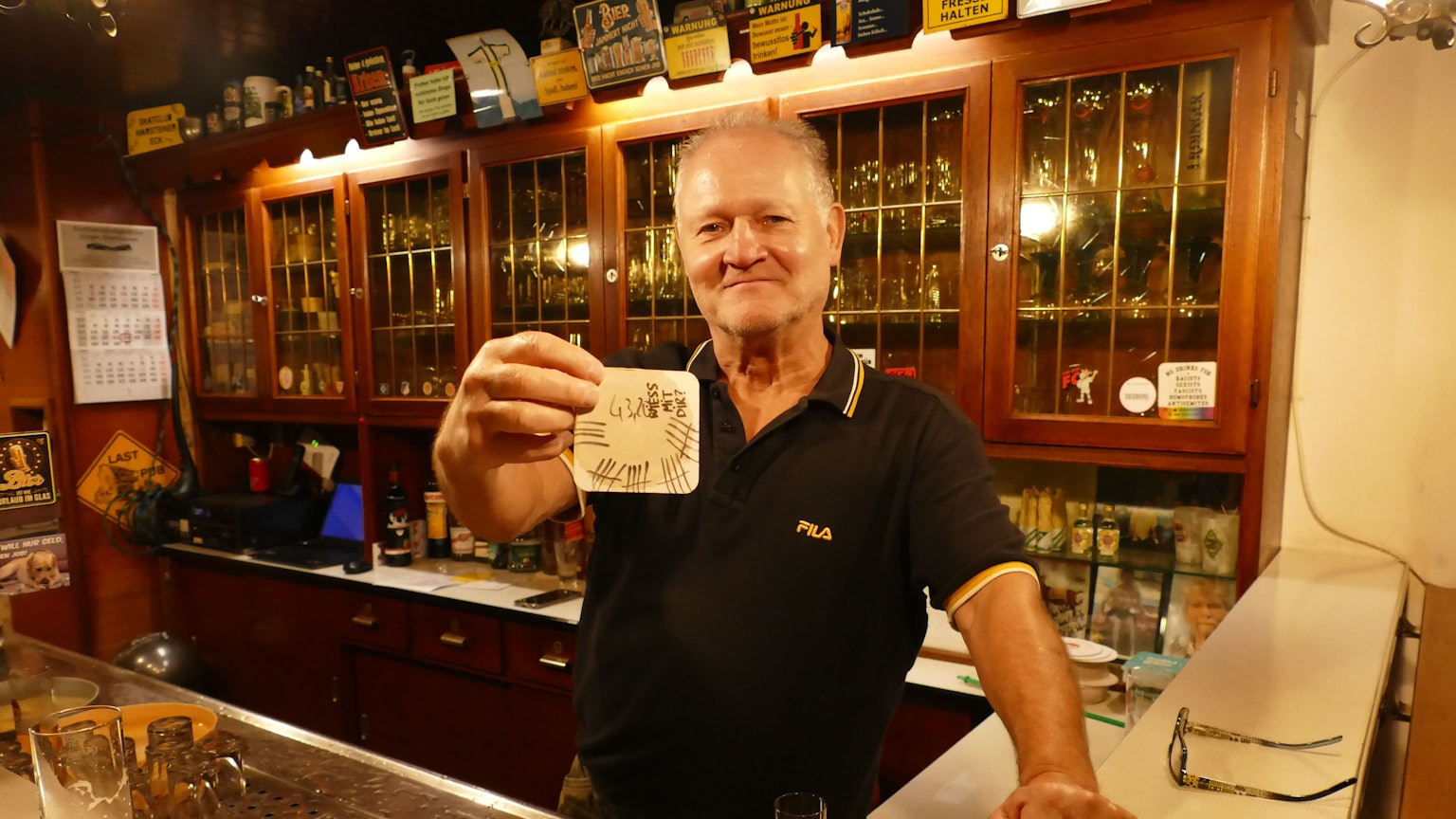 Wolfgang Ort hält einen voll beschriebenen Bierdeckel in der Hand. Er steht hinter der Theke seiner Kneipe.