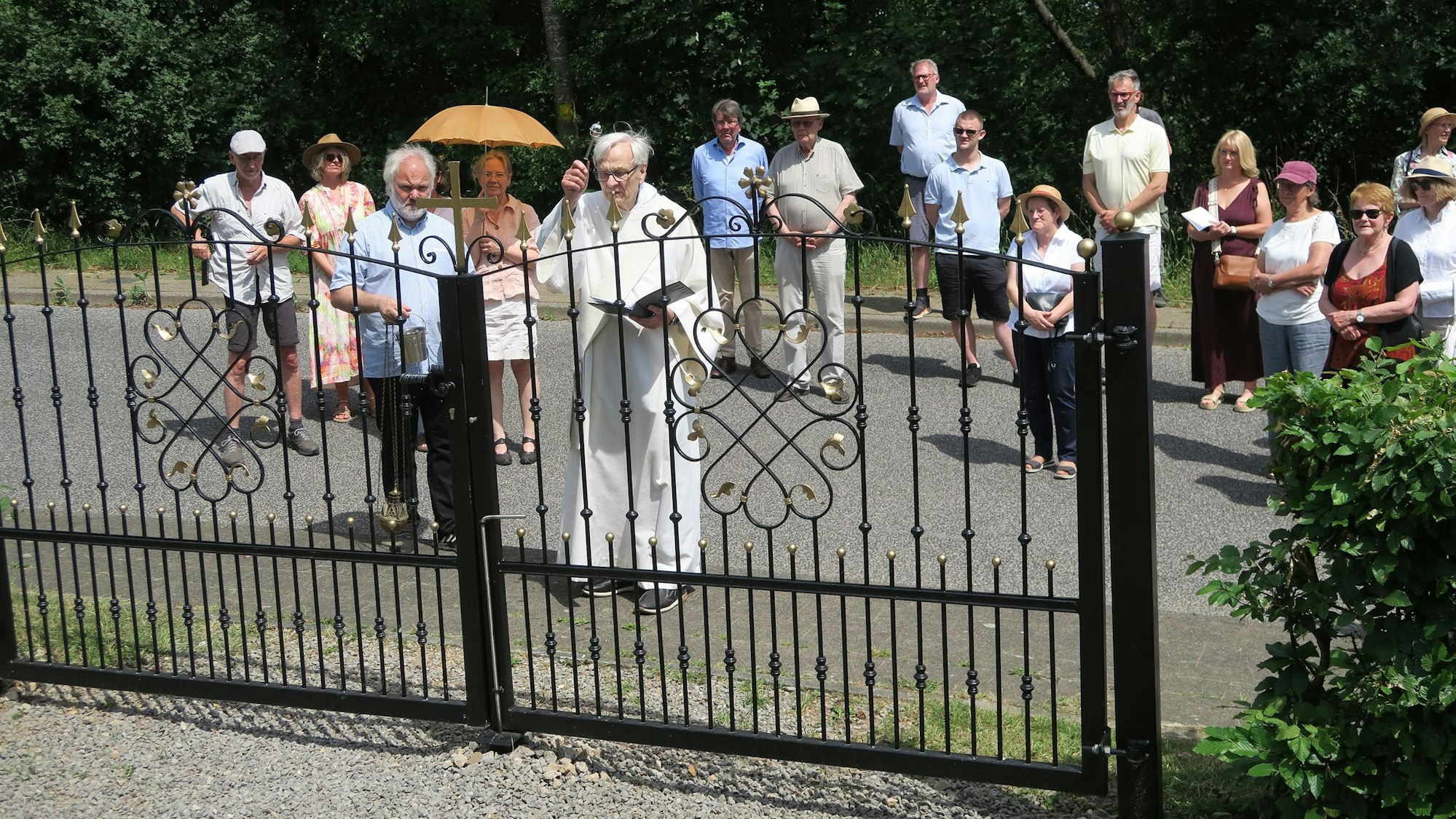 Hinter dem Friedhofstor stehen viele Besucher, ein Geistlicher segnet das Tor.