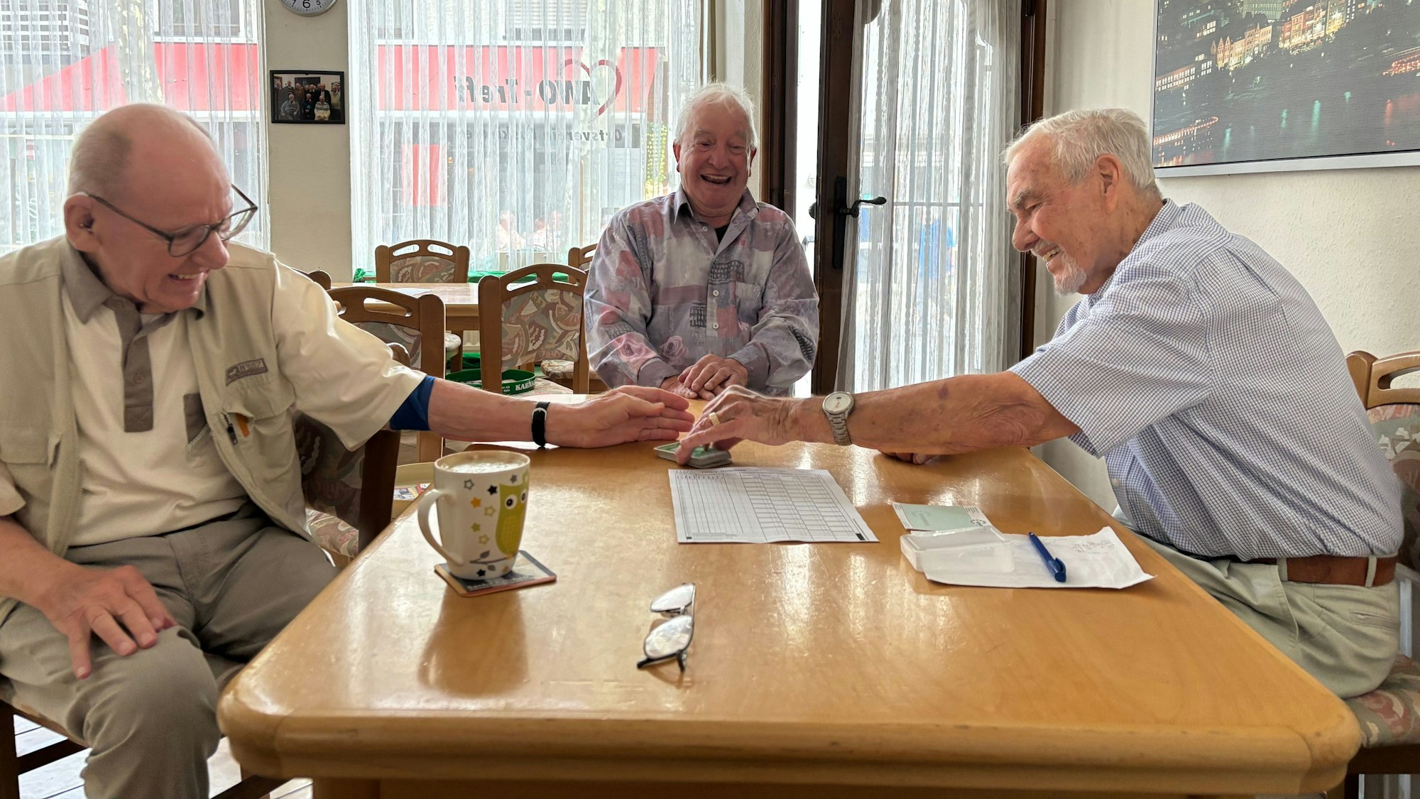 Drei Skatspieler sitzen um einen Tisch und lachen. Einer hat ein Blatt mit einem Kugelschreiber vor sich liegen und greift nach dem Kartenstapel. Ein anderer hat eine Tasse vor sich stehen.