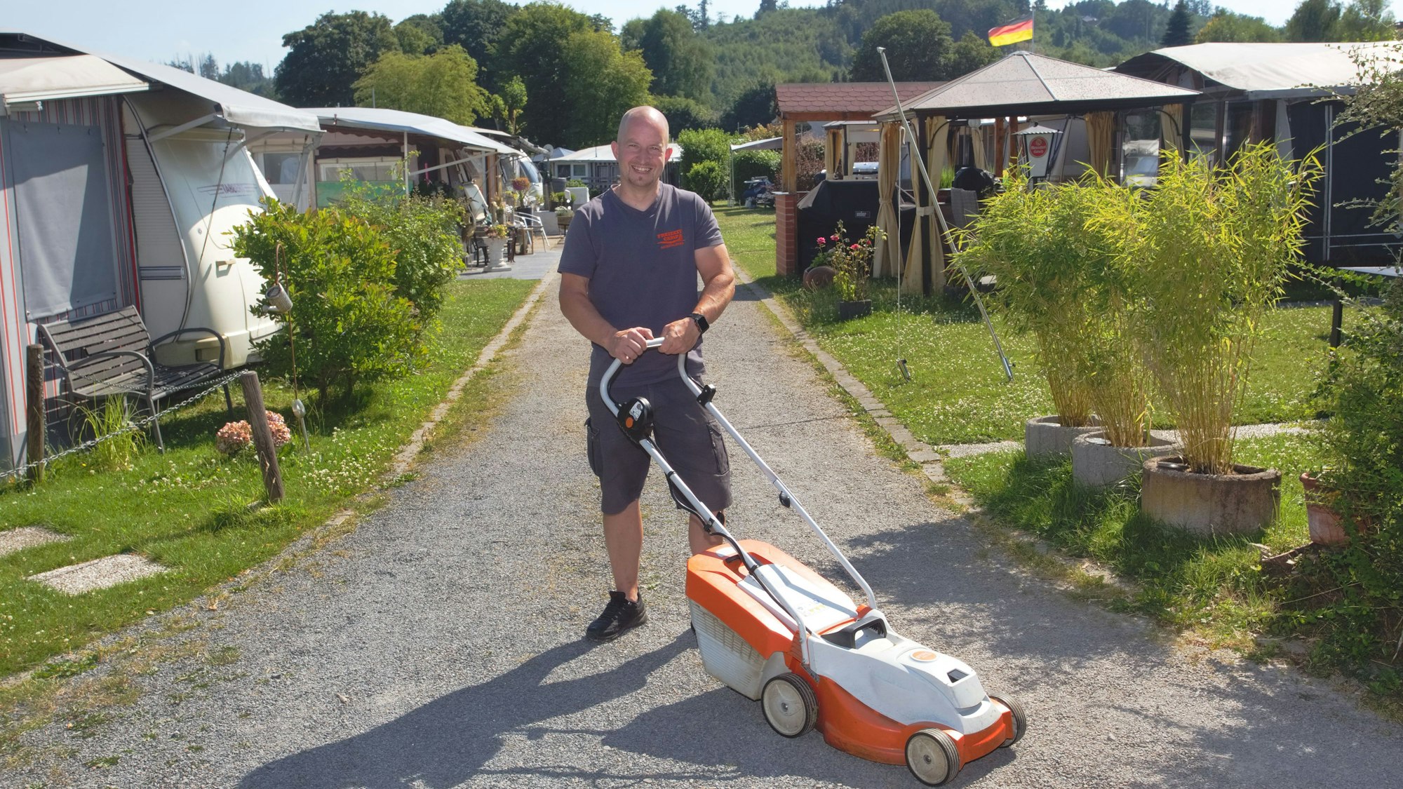 Ein Mann mit Rasenmähre steht auf einem Schotterweg. Neben ihm sind Dauercampingplätze zu sehen.