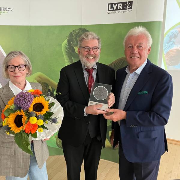 Eine Dame mit Blumenstrauß, zwei Herren mit einer gerahmten Medaille.