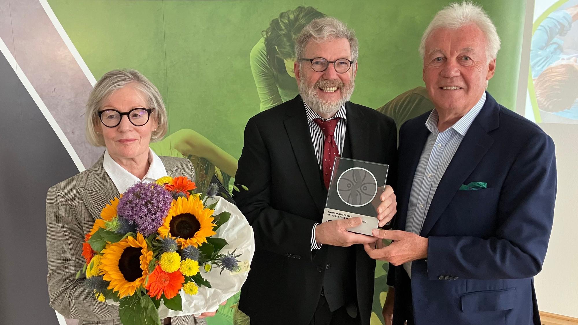 Eine Dame mit Blumenstrauß, zwei Herren mit einer gerahmten Medaille.
