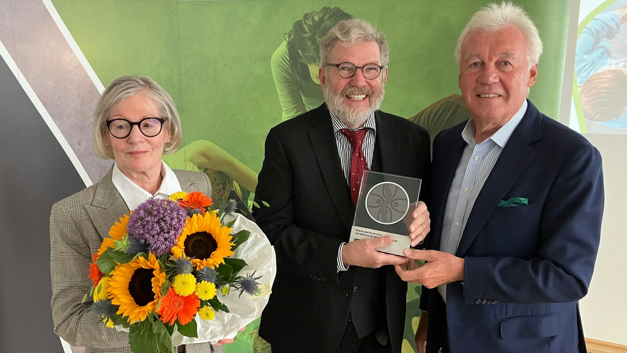 Eine Dame mit Blumenstrauß, zwei Herren mit einer gerahmten Medaille.