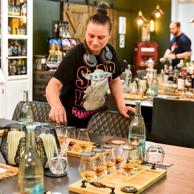 Das Bild zeigt eine Mitarbeiterin bei der Vorbereitung. Auf dem Tisch stehen zahlreiche Gläser mit Whiskey, im Hintergrund ein Schrank voller Flaschen.