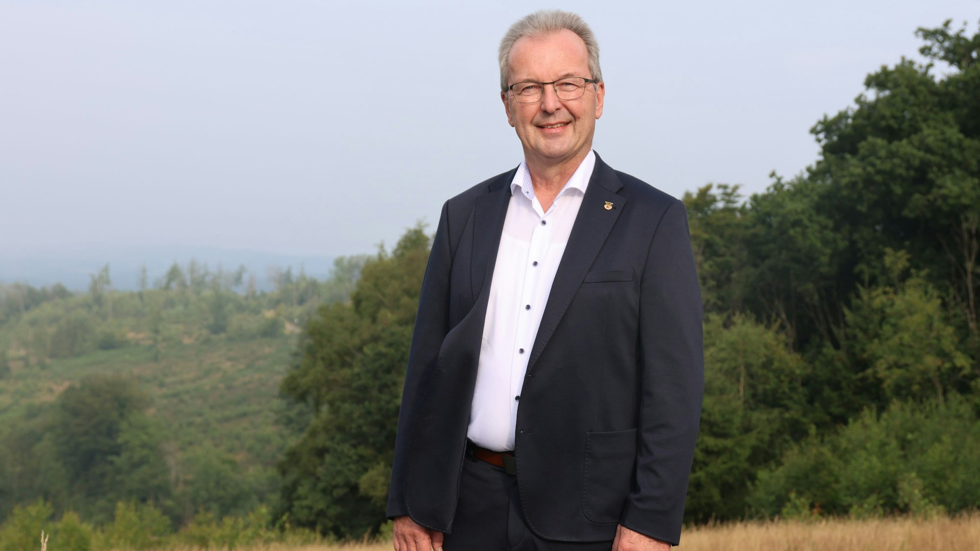 Ein Mann im Anzug steht auf einem Feld mit Bäumen im Hintergrund.