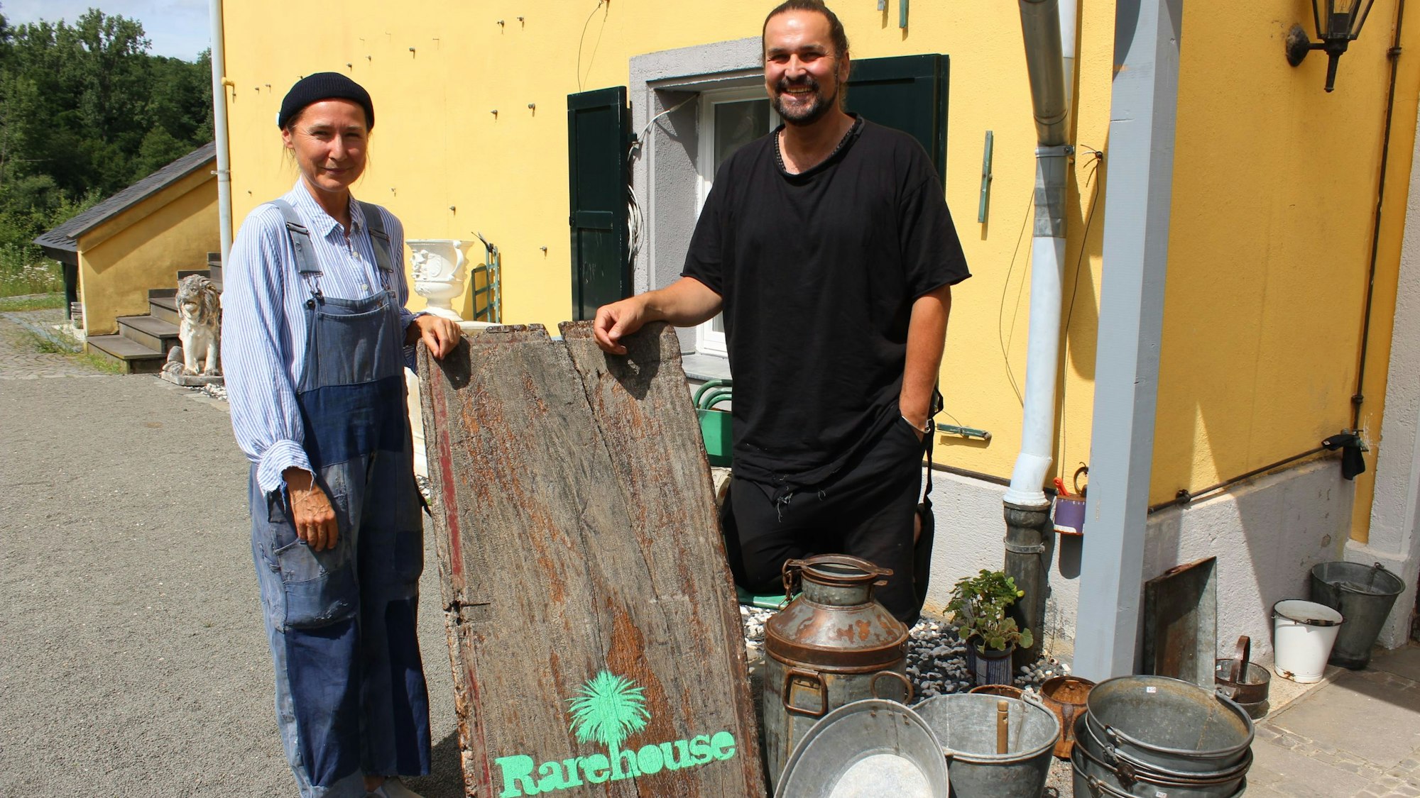 Eine Frau und ein Mann stehen vor einem gelben Haus und halten ein Schild.