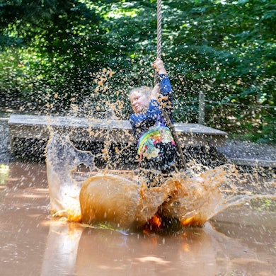Ein Mädchen schwingt sich mit einem Tau über ein Becken mit matschigem Wasser. Mit den Füßen wirbelt es das Wasser auf, das hochspritzt.