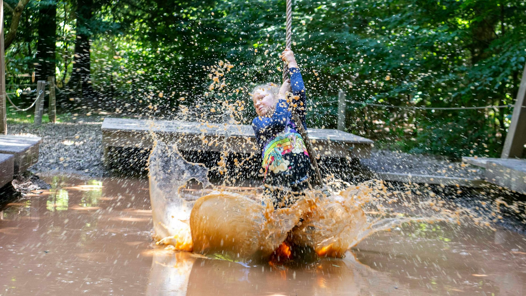 Ein Mädchen schwingt sich mit einem Tau über ein Becken mit matschigem Wasser. Mit den Füßen wirbelt es das Wasser auf, das hochspritzt.