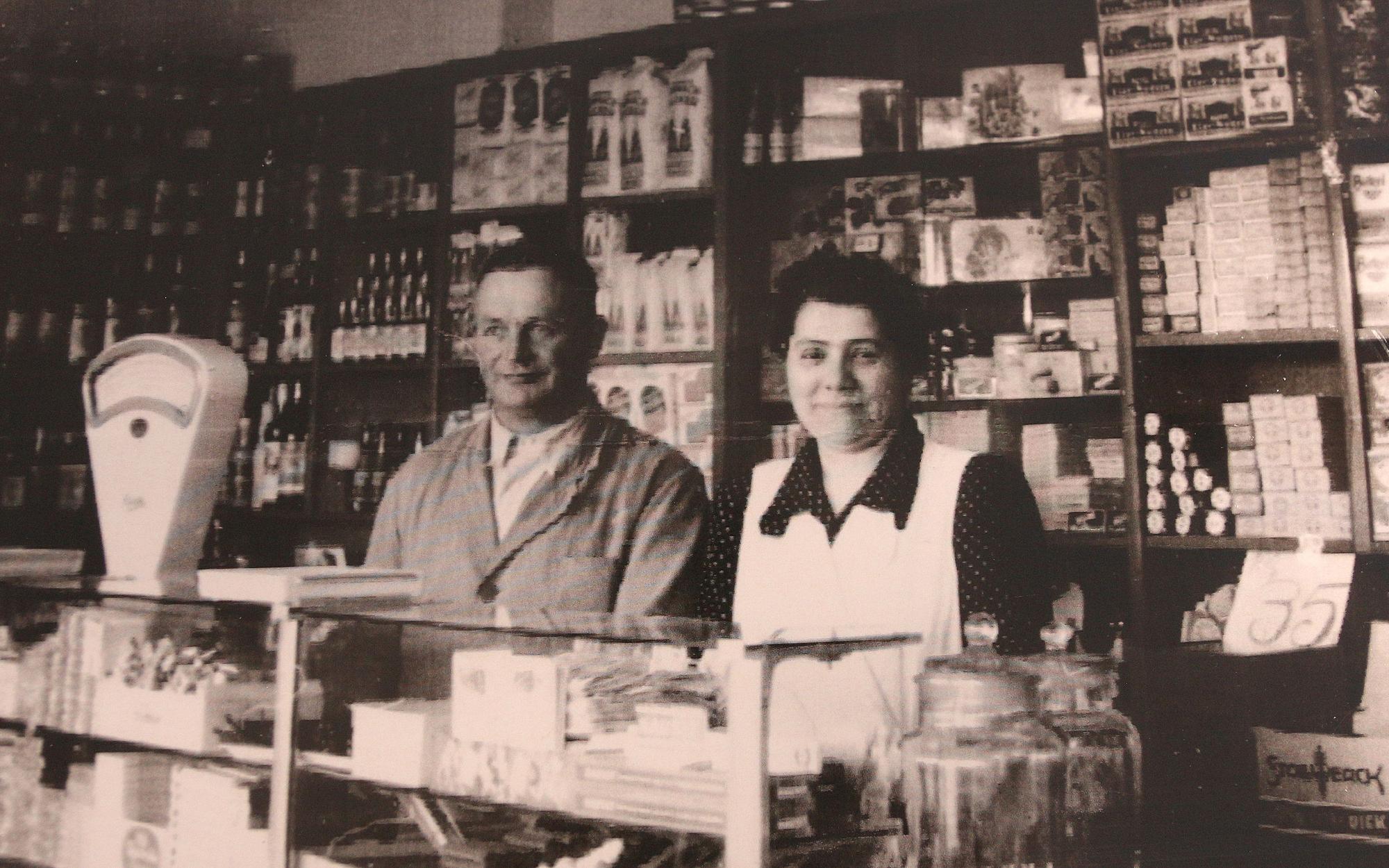 Das Bild zeigt den Laden im Jahr 1930. Das Bild ist schwarz-weiß. Das Ehepaar steht an der Kasse, hinter ihnen viele Waren in Schränken.